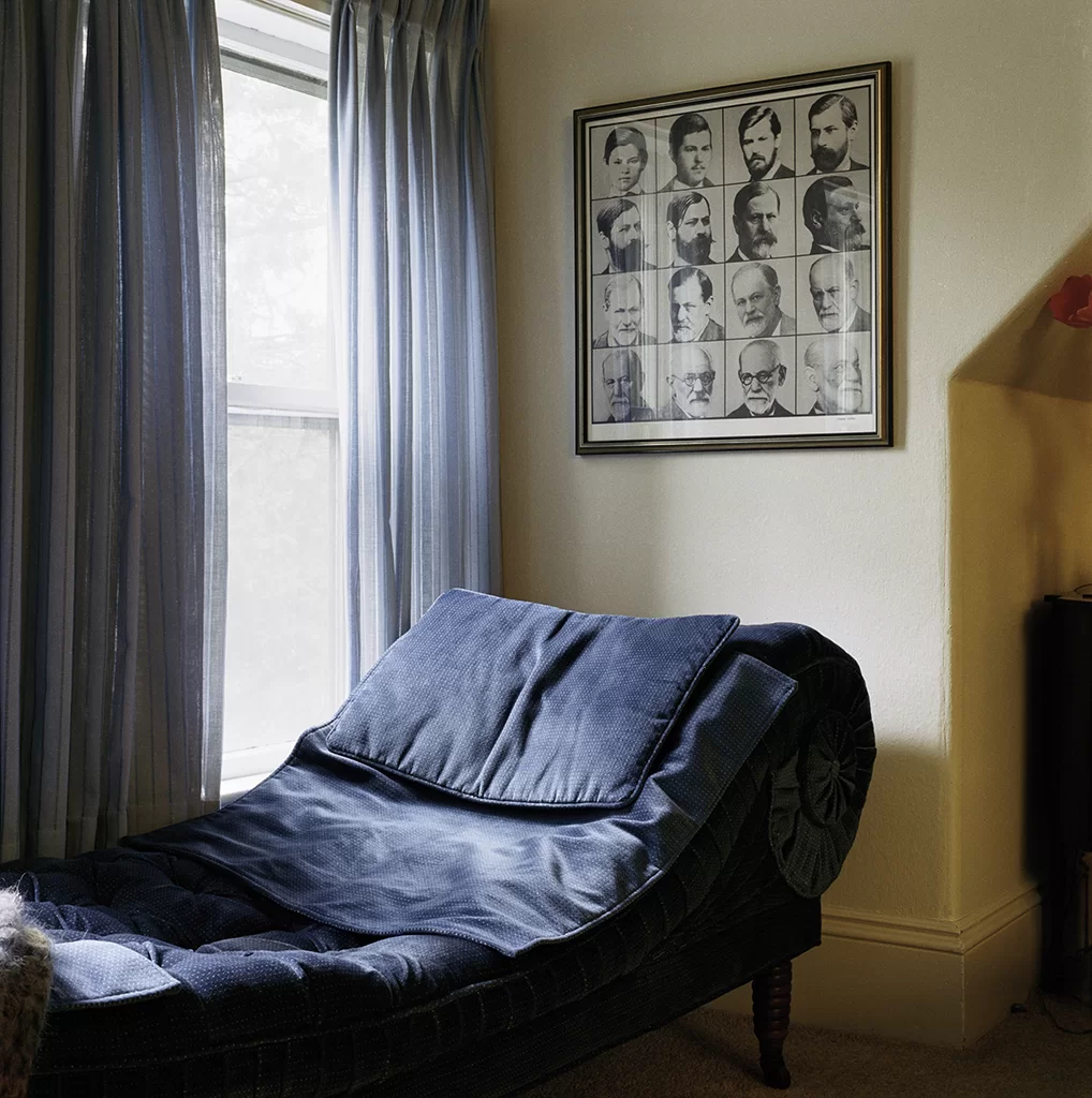 A room with a blue couch and several portraits hanging overhead, next to a window.