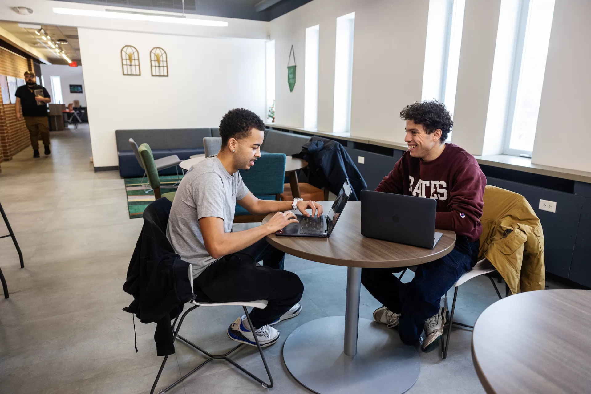 Two men smile and talk while sitting at a table with laptops.