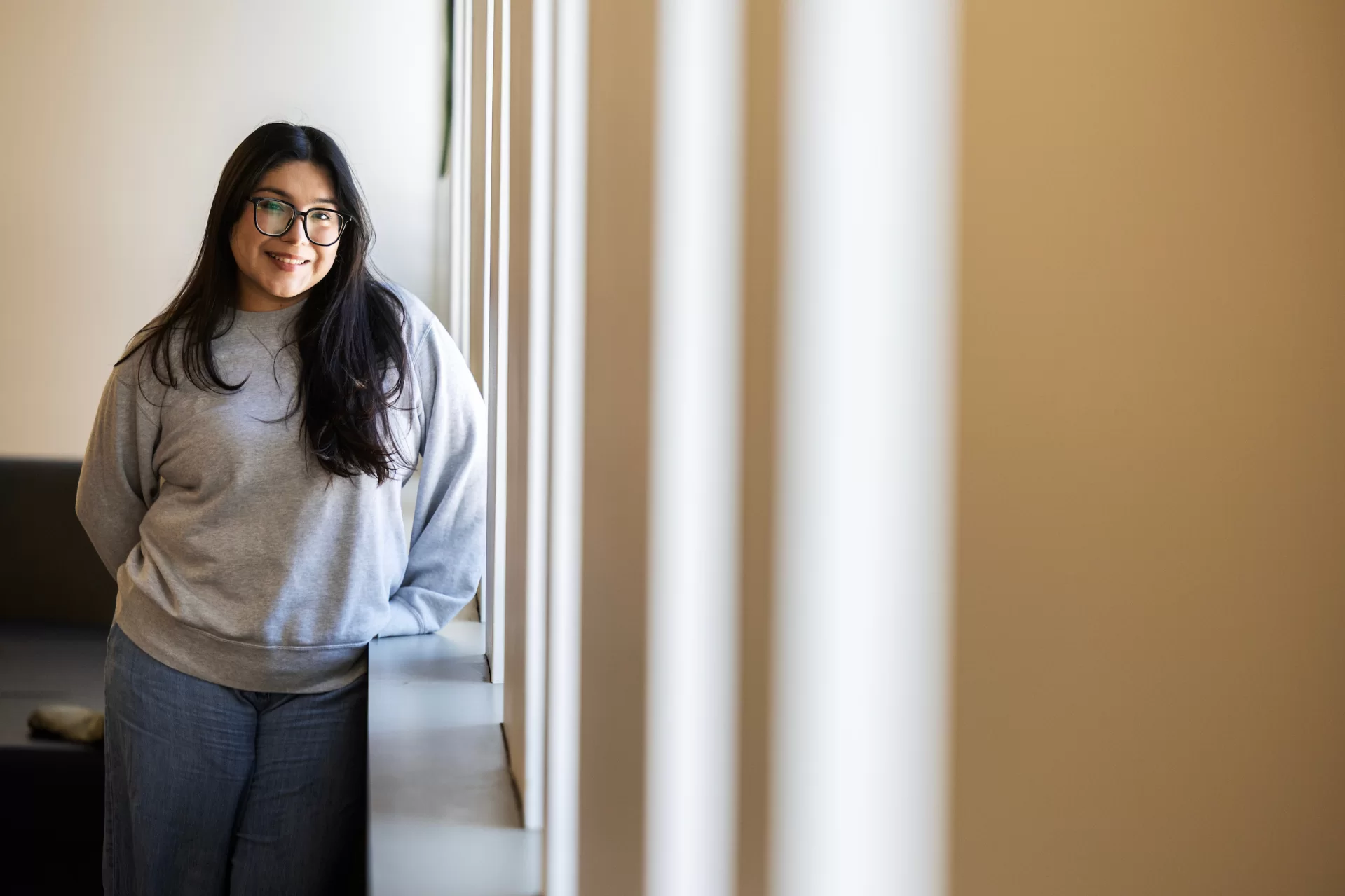 A woman smiles while leaning against a wall lined with windows.