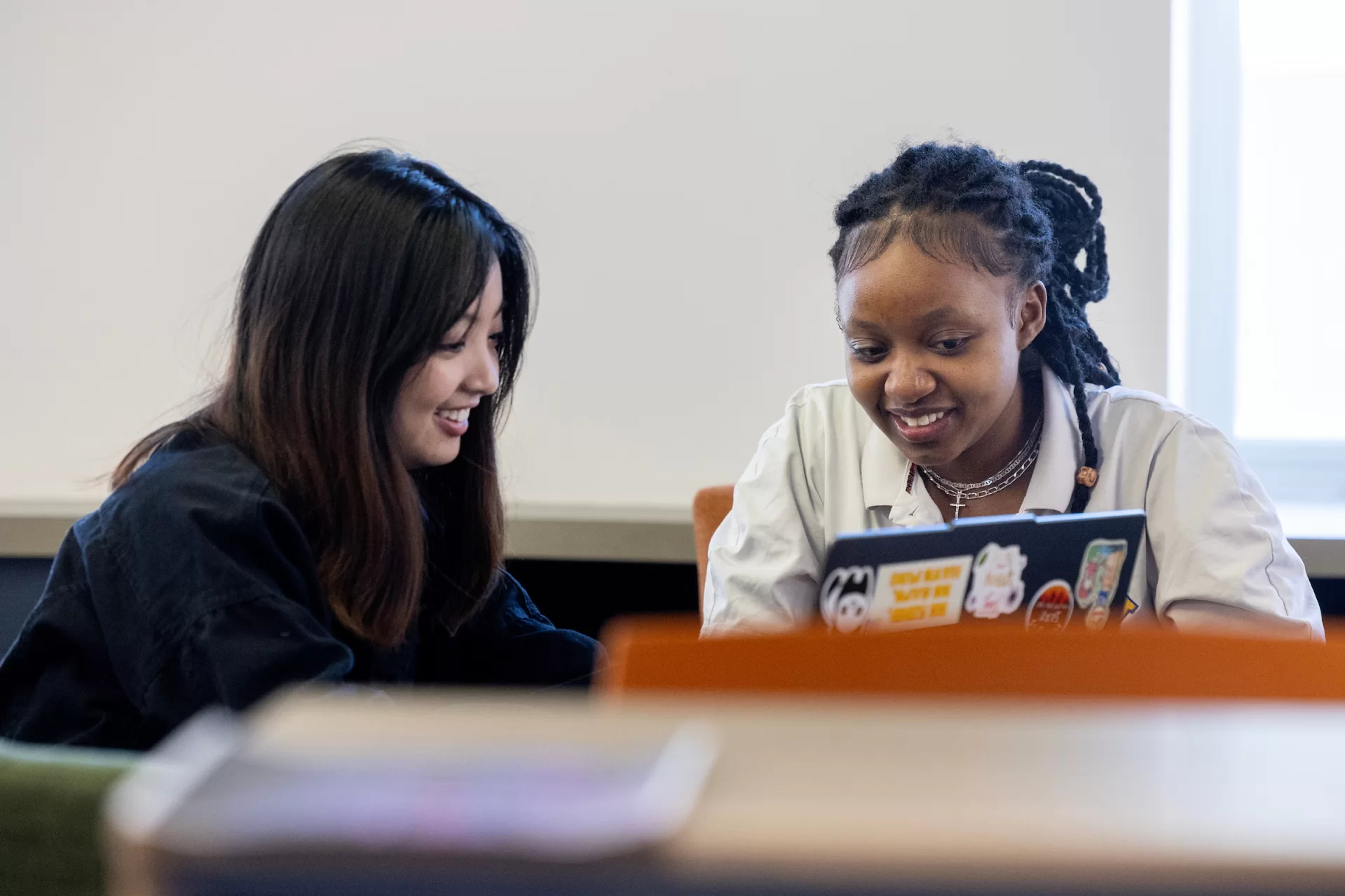 Two women sit at a table with a computer, smiling and talking to each other.