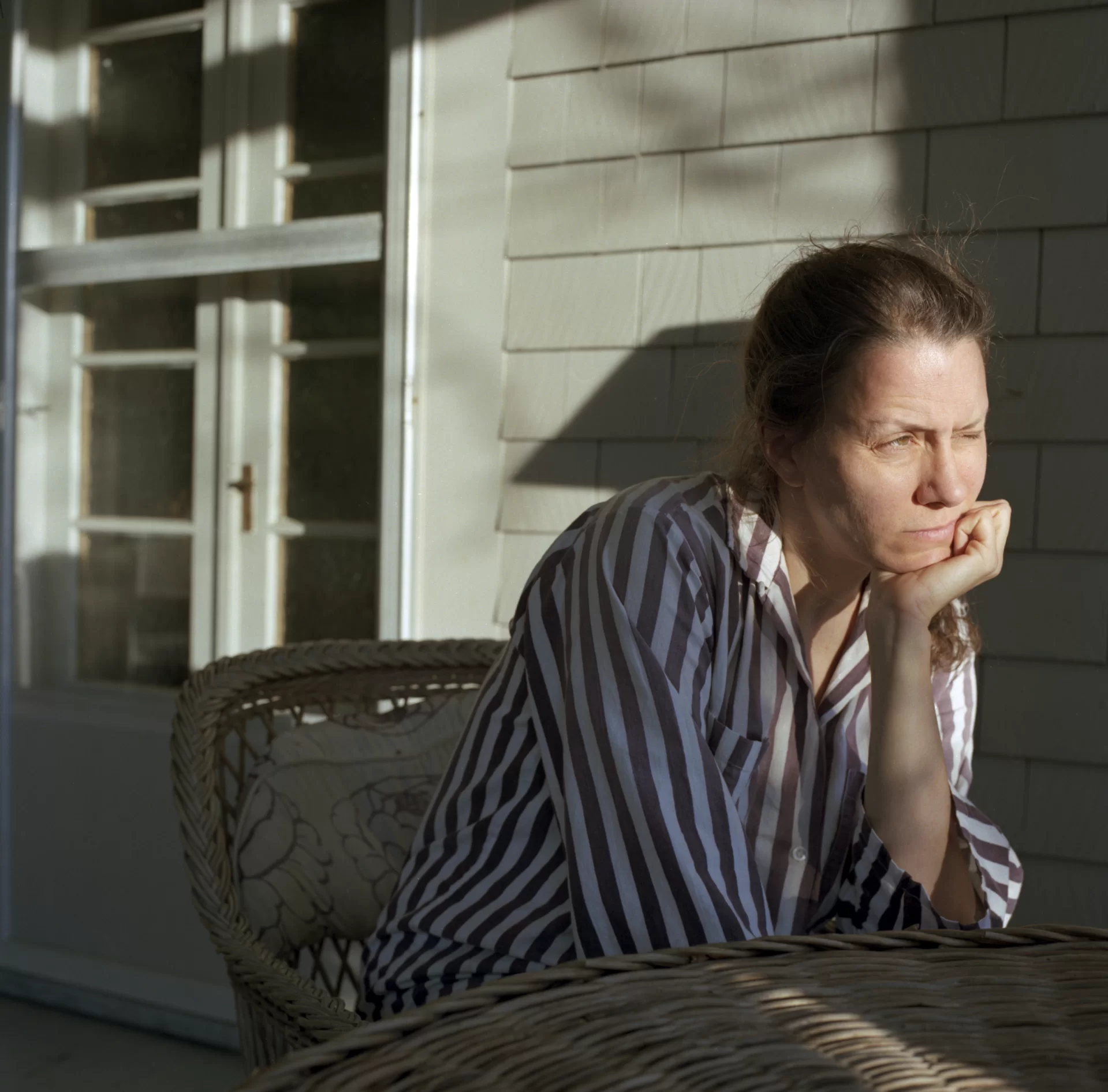 A woman sits on a sunny porch, squinting and gazing outward.