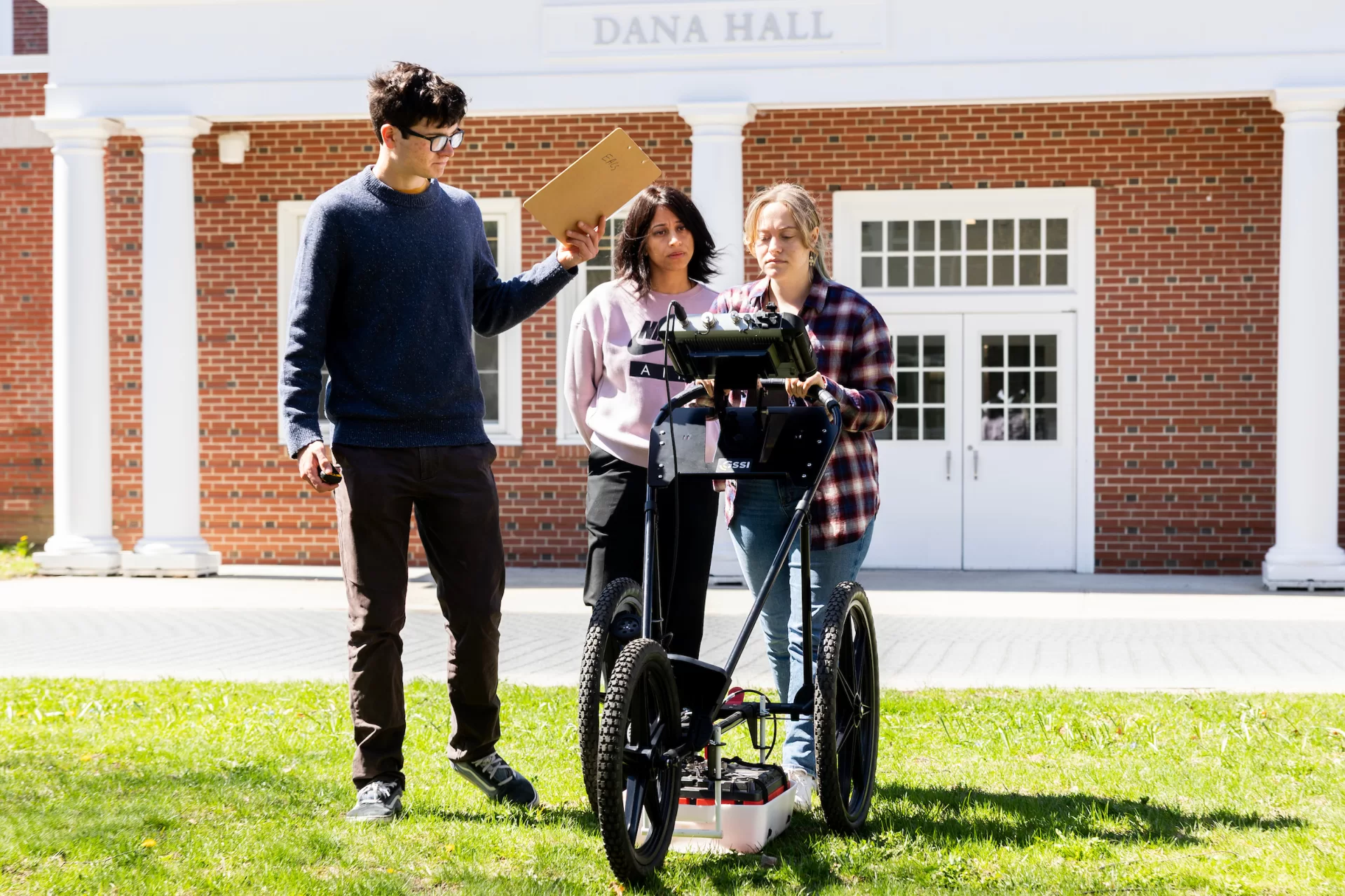 Three people push a large scientific device across a college campus.