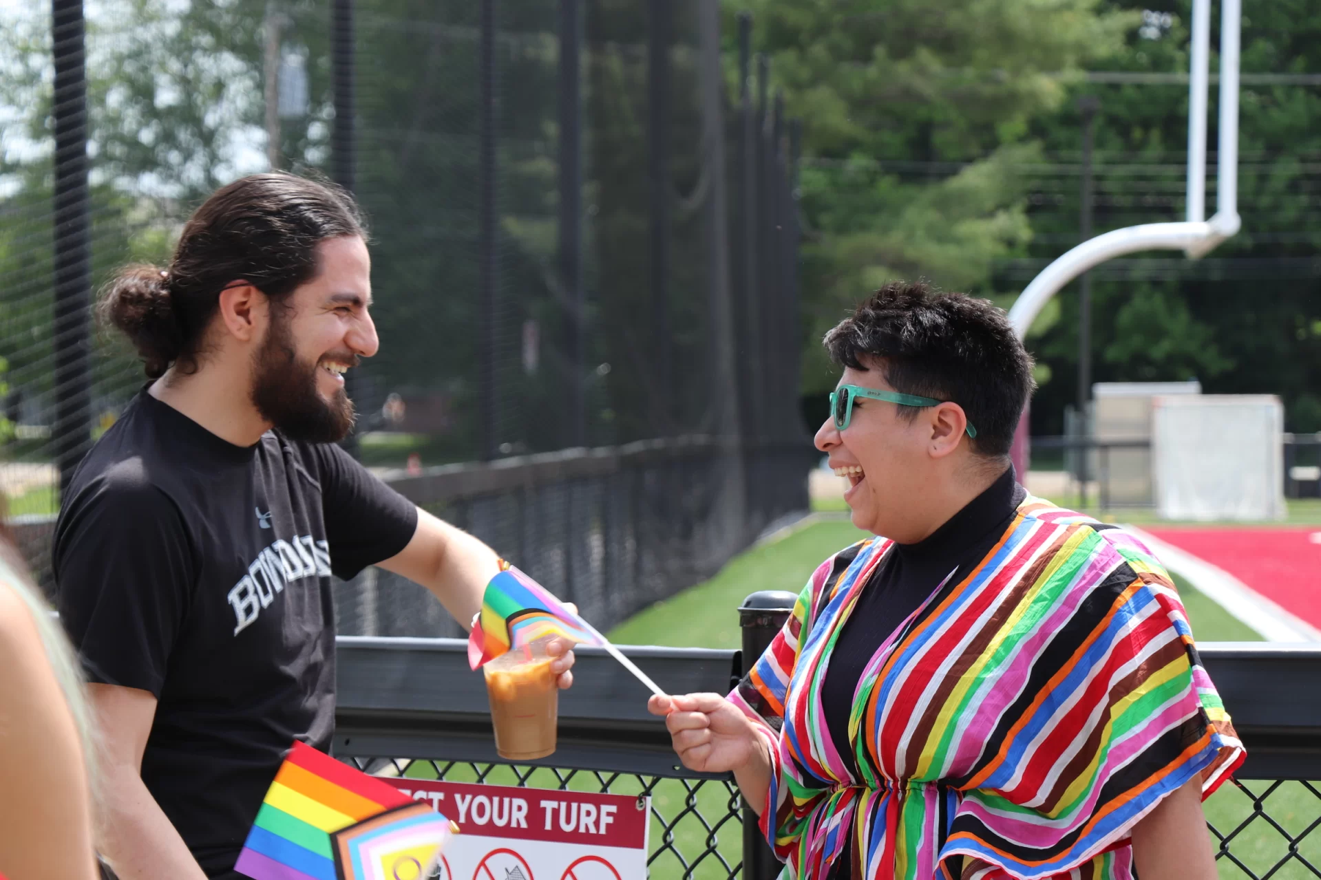 Two people dressed in Pride clothing lean on a fence and talk
