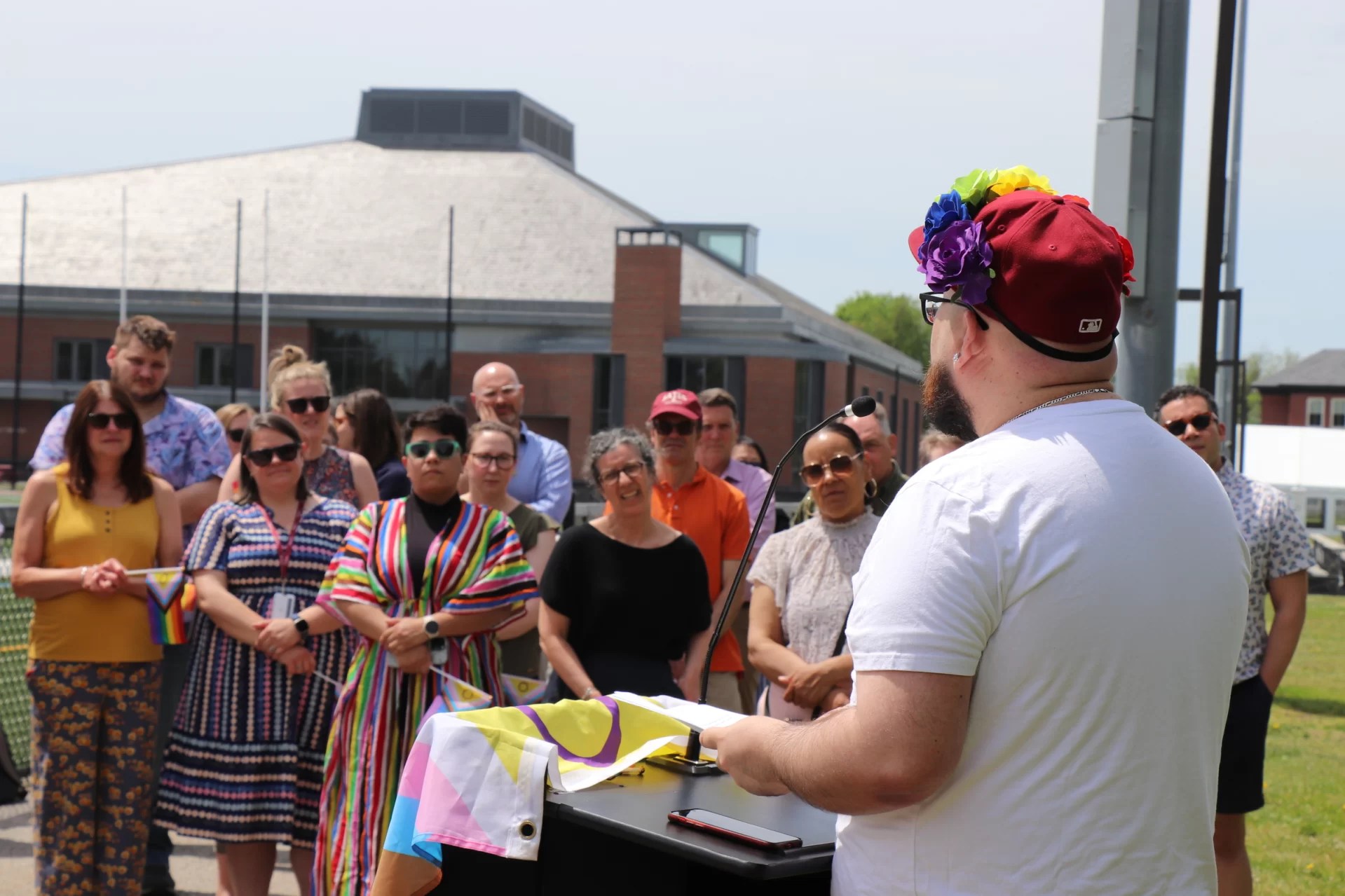 A person wearing a rainbow headband delivers  a speech to a crowd wearing Pride clothing