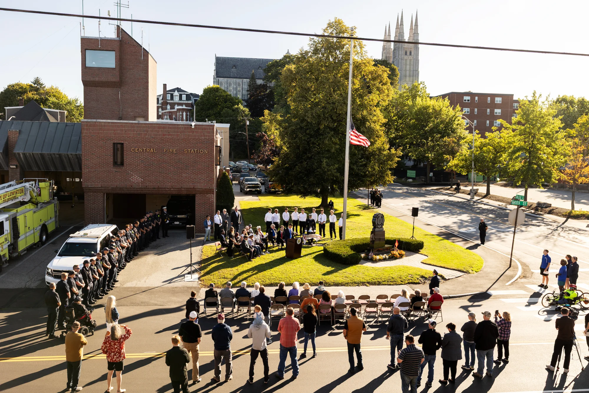 From above, a group of people, including firemen and a cappella singers in white T-shirts, gather outside of a firehouse, some sitting, some standing