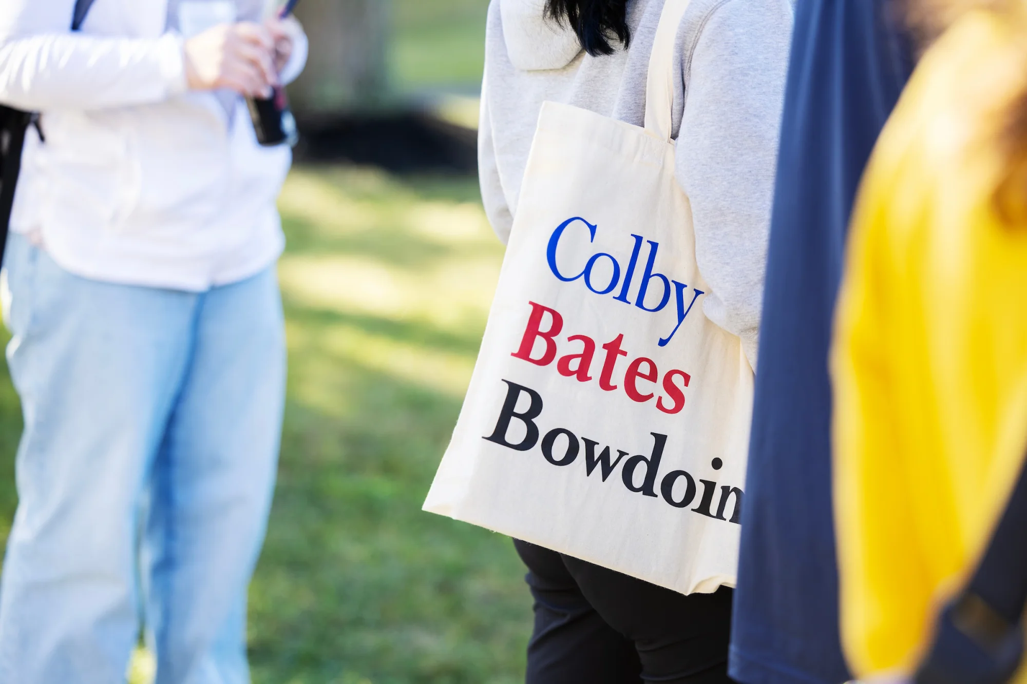  A tote bag over a person's shoulder that says "Colby Bates Bowdoin" in blue, red, and black lettering