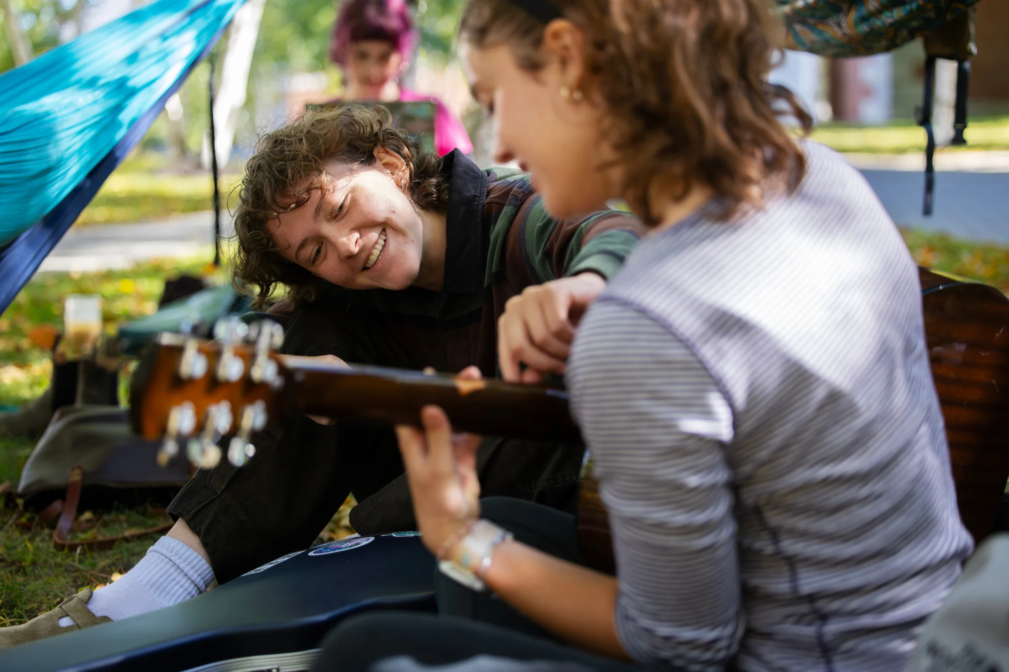 A person leans forward to adjust another person's finger placement on an acoustic guitar. They are sitting outside, with a hammock in the background.