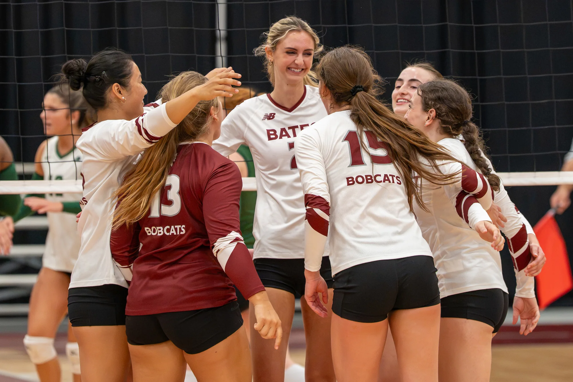 A group of volleyball players cheer and embrace on the court, with the net in the background