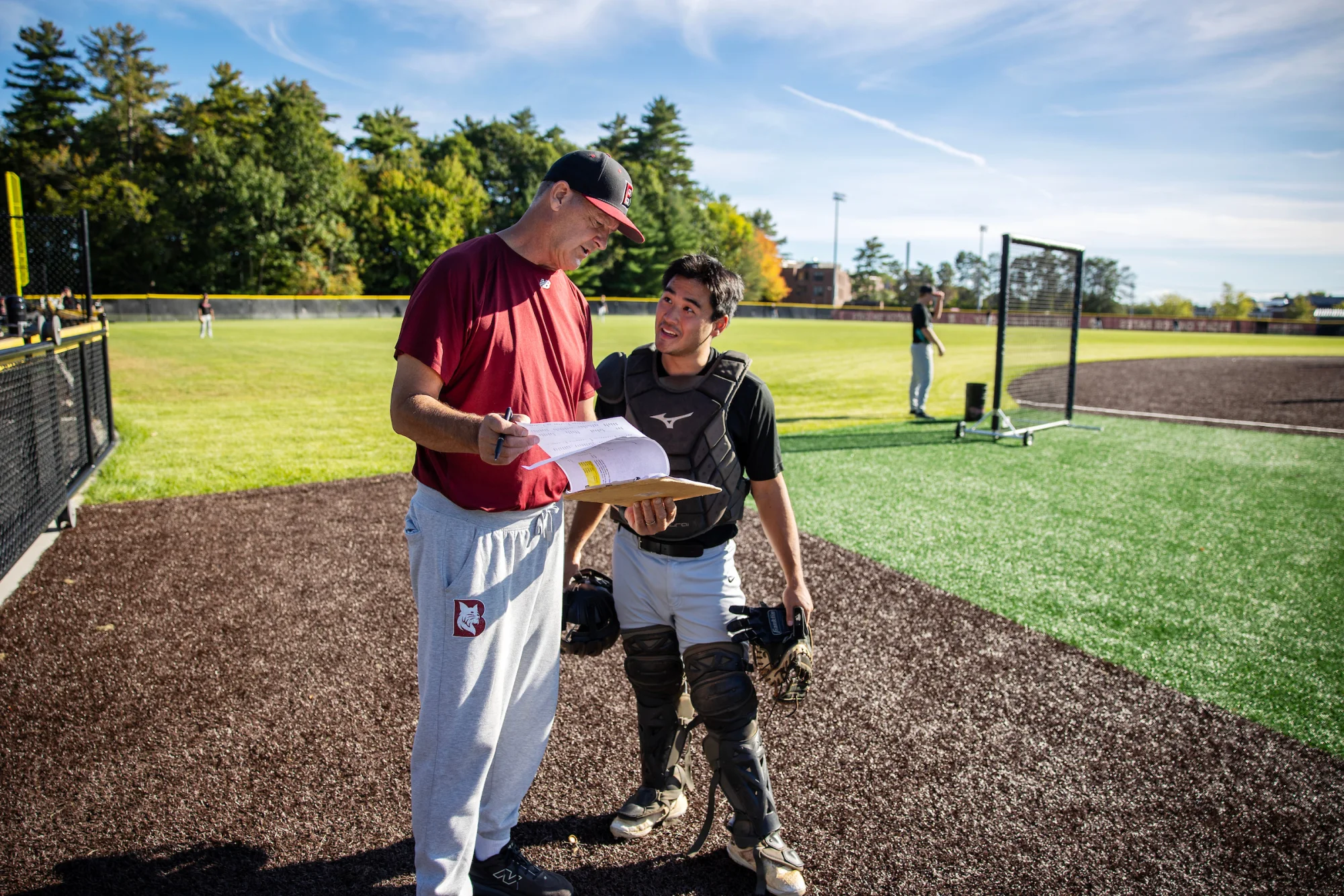 Two people, one in baseball gear, stand chatting on the side of a baseball diamond