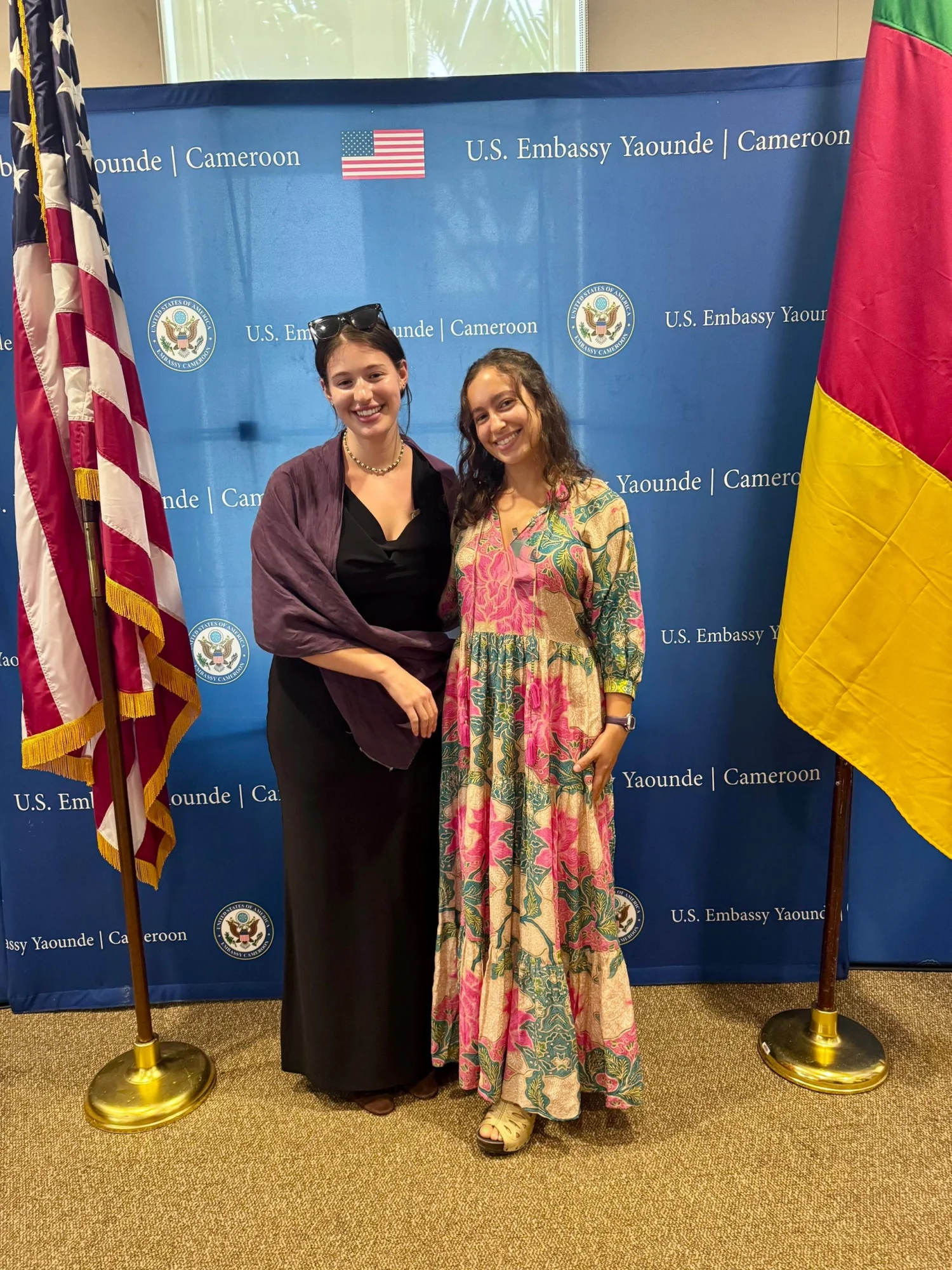 Two women stand smiling in front of a background that says "U.S. Embassy Yaounde | Cameroon," flanked by flags from the U.S. and Cameroon.