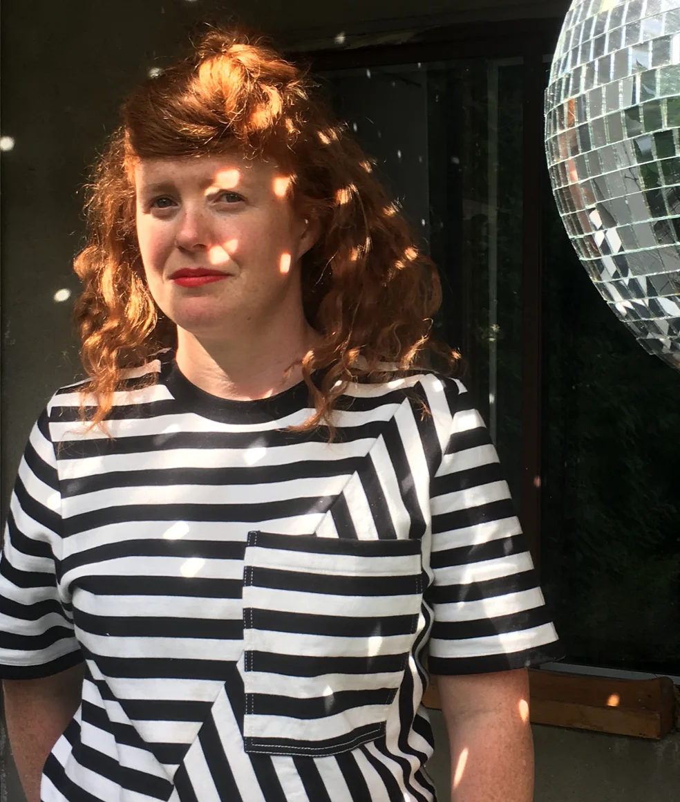 A woman with shoulder-length, curly red hair wearing a black and white striped patterned shirt. There is a mirrored disco ball in the top right hand corner of the photograph. Bright circles of light reflect from the disco ball onto the woman face and torso.