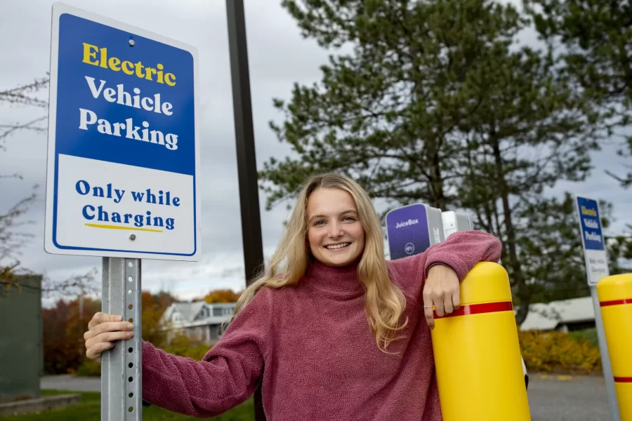 Ecorep Tamsin Stringer ’22 of Bloomington, Ind., poses at the new electric charger stations behind Underhill in the Merrill Gym parking lot.

“Underhill Electric Vehicle Chargers Project “
Bates has installed other EV chargers in the past. This project will be different for three primary reasons. First, we have already received a grant from CMP for the make-ready infrastructure portion of the project, which has historically been the bulk of the expenses for EV charger installations. Secondly, this project will include installing level 2 chargers for the first time at Bates, which will allow for monetary collections for charging, tiered charging for different kinds of customers, and incentivize to move one’s car once it's fully charged. Finally, this project allows for future EV charger installations in the same location for much lower cost, because the make-ready infrastructure for more EV chargers will be easily accessible.